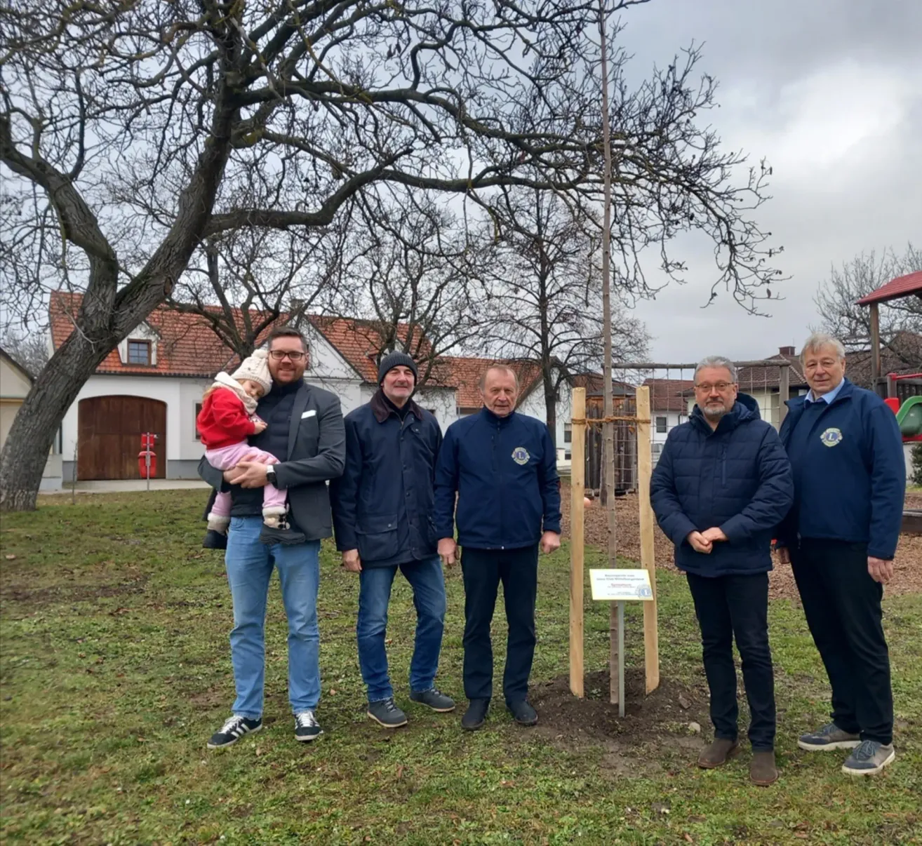 Fünf Männer in passenden Mänteln stehen um eine neu gepflanzte Baum herum. Ein Mann hält ein Baby in den Armen. Hinter ihnen sind ein Haus und Bäume zu sehen.
