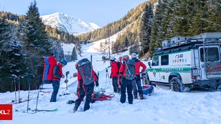 Eine Gruppe von Skifahrern und Rettern steht in tiefem Schnee, trägt Helme und Rucksäcke. Sie befinden sich in der Nähe eines schneebedeckten Berges mit Bäumen. Ein Fahrzeug mit der Aufschrift 'Bergrettung' ist in der Nähe geparkt.