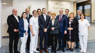 Eine Gruppe von formell gekleideten Menschen, darunter medizinisches Personal, steht in einem Raum zusammen. Hinter ihnen befindet sich ein Schild mit der Aufschrift Herzlich Willkommen.