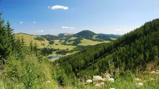 Ein Panoramablick auf eine bergige Region mit sanften Hügeln, einem See und üppigen grünen Feldern unter einem klaren blauen Himmel.