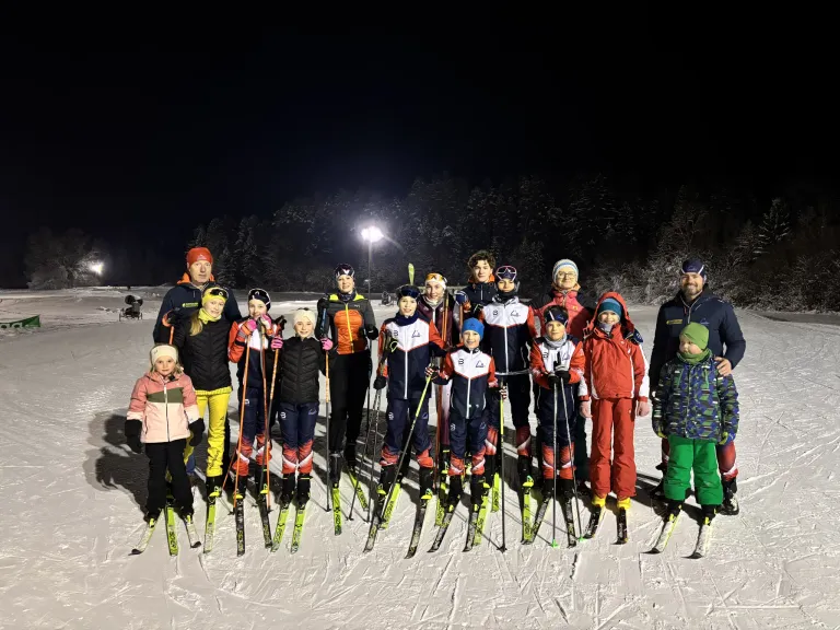 Eine Gruppe von Skifahrern steht bei Nacht auf einem verschneiten Feld und trägt verschiedene Winterkleidung. Hinter ihnen sind Bäume und eine beleuchtete Laterne.