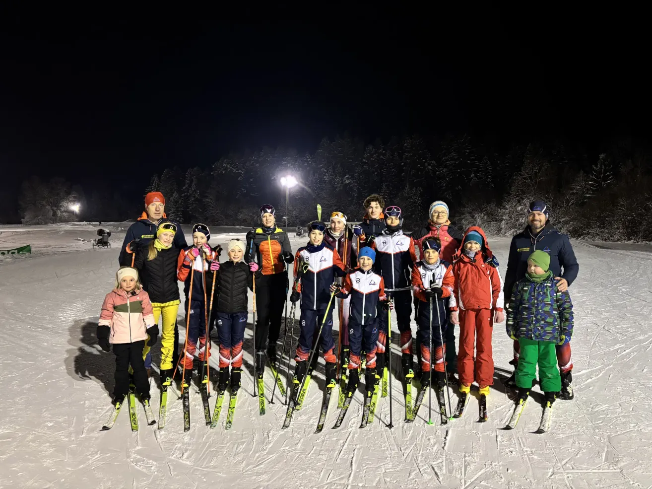 Eine Gruppe von Skifahrern steht bei Nacht auf einem verschneiten Feld und trägt verschiedene Winterkleidung. Hinter ihnen sind Bäume und eine beleuchtete Laterne.