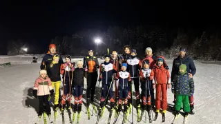 Eine Gruppe von Skifahrern steht bei Nacht auf einem verschneiten Feld und trägt verschiedene Winterkleidung. Hinter ihnen sind Bäume und eine beleuchtete Laterne.