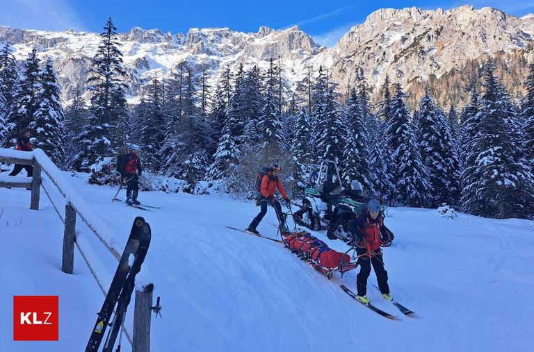 Vier Personen skifahren auf einem verschneiten Berg mit einem Schlitten vor ihnen, der jemanden Verletzten trägt. Bäume bedecken den Berghang.