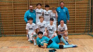 Eine Fußballmannschaft mit weißen Trikots posiert für ein Foto mit zwei Trainern vor einer Holzwand. Ein Junge hält eine Trophäe, während er auf dem Boden liegt.