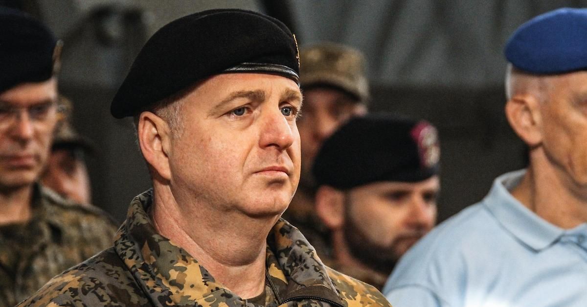 Ein Militäroffizier in Tarnuniform steht aufmerksam da, mit ernster Miene nach vorne gerichtet. Dahinter ist ein weiterer Offizier in ähnlicher Kleidung zu sehen, sowie ein Mann in einem blauen Hemd.