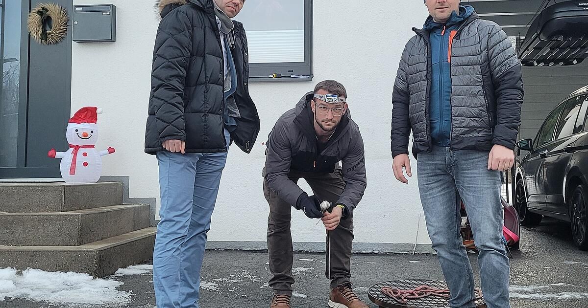 Drei Männer, die warm gekleidet sind, stehen draußen vor einem Gebäude. Ein Mann hockt sich hin, hält ein Werkzeug, während die anderen beiden hinter ihm stehen.