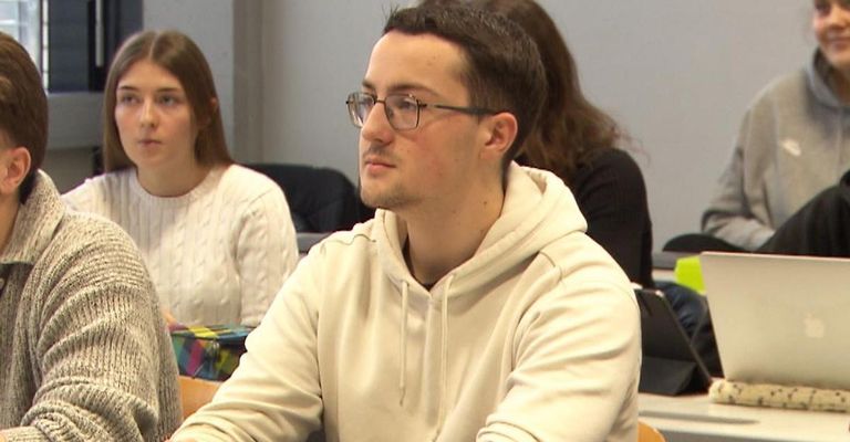A man in glasses and a hoodie sits in a classroom, looking forward with a serious expression. Behind him, a woman in a white sweater is seated. A woman in black is seated behind them.