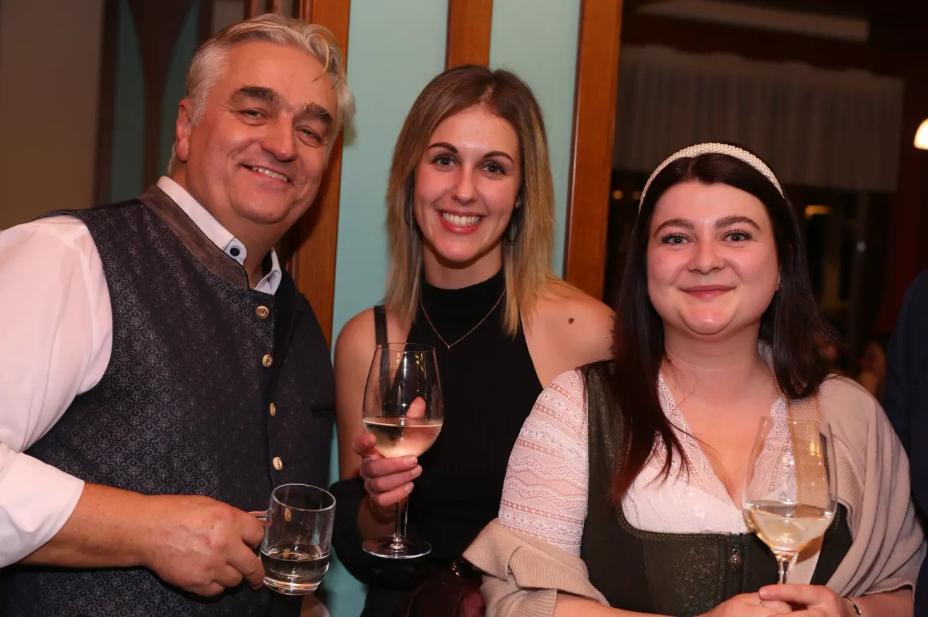 Drei Menschen lächeln für ein Foto. Ein Mann links, eine Frau in der Mitte mit einem Weinglas und eine Frau rechts mit einem Stirnband. Sie tragen alle festliche Kleidung.