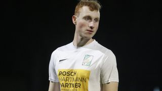 Ein junger Mann mit roten Haaren und blauen Augen trägt ein weißes Shirt mit einem gelben Aufnäher und einem grünen Aufnäher auf der linken Seite. Das Shirt trägt die Aufschrift 'POSCH HARTMMANN PARTNER'. Der Hintergrund ist dunkel.