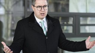 Ein Mann in einem schwarzen Mantel, einer Brille und einer grün gestreiften Krawatte steht und spricht in einem Gebäude mit Glaswänden.