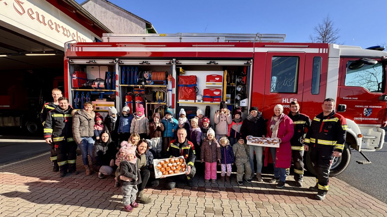 Eine Gruppe von Menschen, darunter Kinder, posiert für ein Foto vor einem Feuerwehrauto mit offener Tür. Sie halten Tabletts mit Donuts.