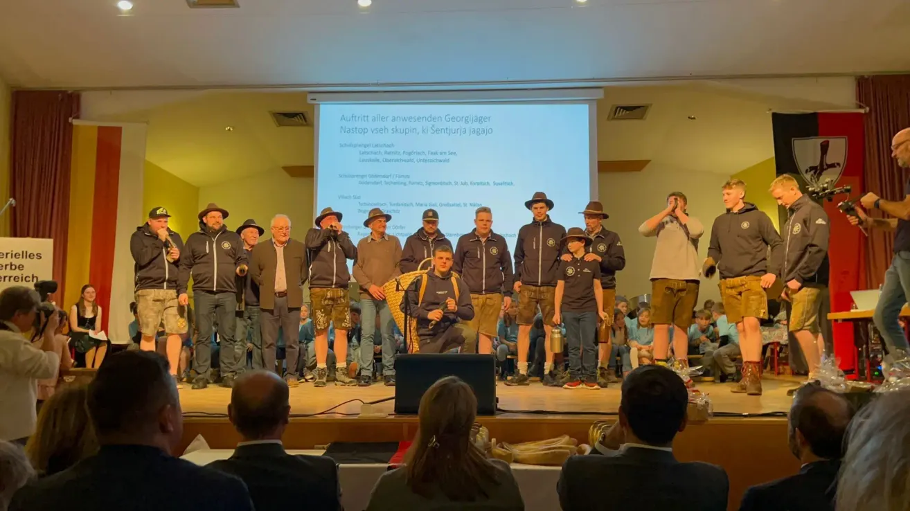 Eine Gruppe von Leuten in Jacken und Mützen steht auf der Bühne und blickt auf eine Projektionsleinwand. Die Bühne befindet sich vor einem sitzenden Publikum. Auf der Bühne steht ein Lautsprecher.