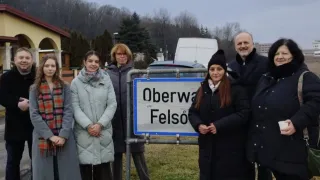 Eine Gruppe von Personen in Winterkleidung steht um ein Straßenschild für Oberwald Felso im Freien. Im Hintergrund sind Bäume und ein Gebäude zu sehen.