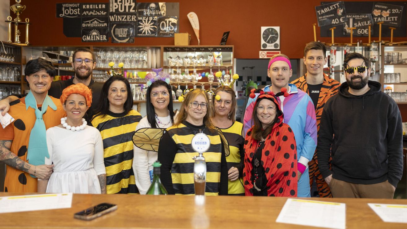 Eine Gruppe von Erwachsenen in thematischen Kostümen posiert für ein Foto in einer Bar mit einer roten Wand und verschiedenen ausgestellten Gegenständen.
