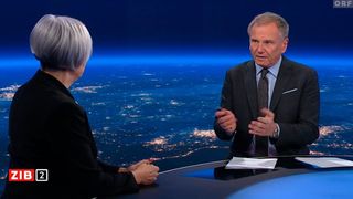 Ein älterer Mann spricht in einem Nachrichtenstudio und gestikuliert mit den Händen. Eine Frau hört aufmerksam zu. Im Hintergrund ist eine Stadtlandschaft mit Lichtern zu sehen.