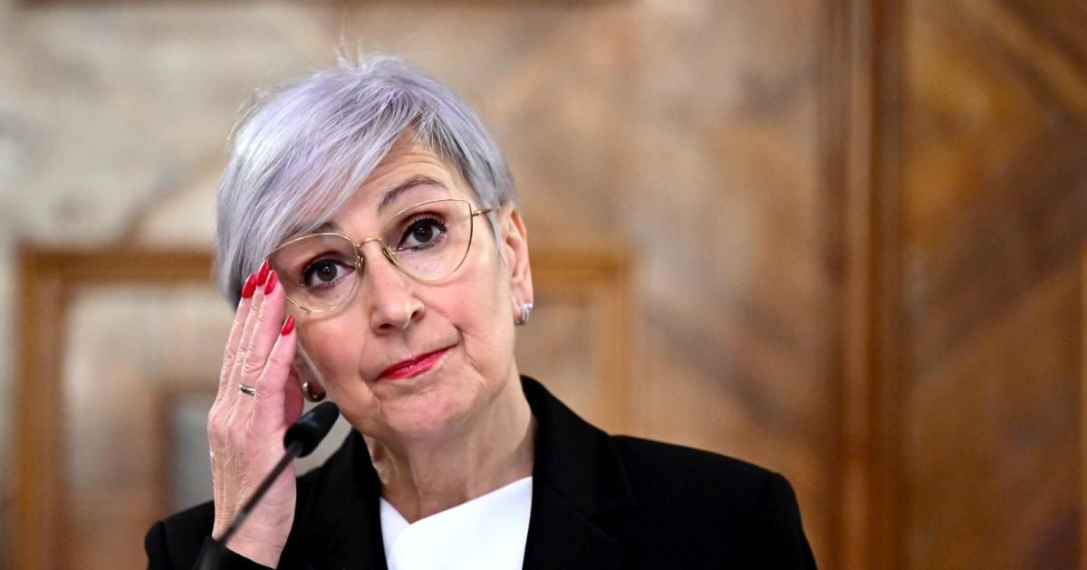 Eine ältere Frau mit grauem Haar und Brille spricht in ein Mikrofon, ihre Hand ruht auf ihrer Wange.