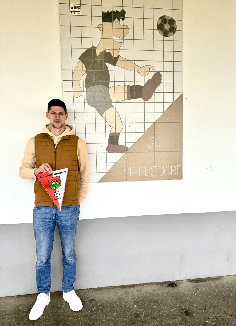 Ein Mann steht vor einer Wand mit einer großen Zeichnung eines Fußballspielers und hält eine dreieckige Flagge mit dem Text S.C.Mariasdorf. Er trägt eine braune Weste und blaue Jeans.