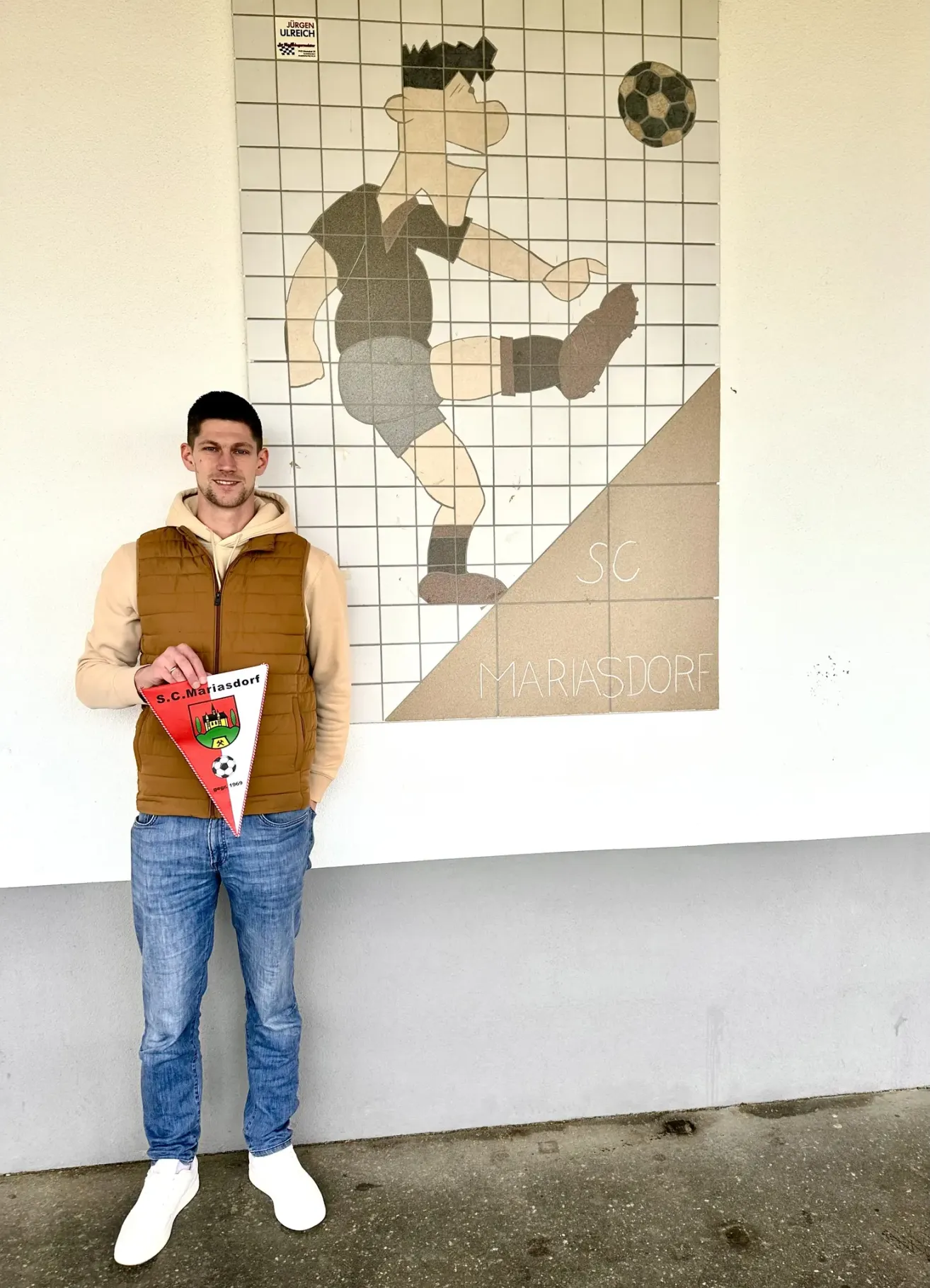 Ein Mann steht vor einer Wand mit einer großen Zeichnung eines Fußballspielers und hält eine dreieckige Flagge mit dem Text S.C.Mariasdorf. Er trägt eine braune Weste und blaue Jeans.