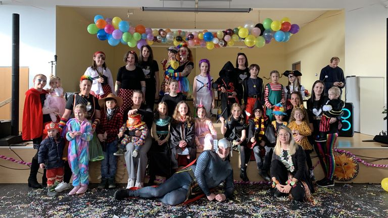 Eine Gruppe von Kindern und Erwachsenen in Kostümen posiert für ein Foto in einem dekorierten Raum mit Luftballons. Ein Mann in einem Superheldenkostüm liegt auf dem Boden.