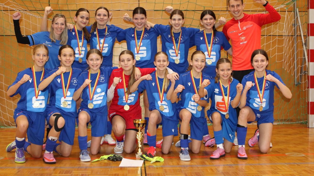 A group of young female athletes wearing blue uniforms and gold medals pose with a trophy on a wooden floor.