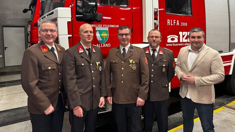Five men in military uniforms are standing in front of a red fire truck, likely in a ceremonial setting.