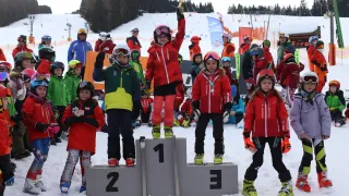 Fünf kleine Kinder stehen auf einem Podest bei einem Skiwettbewerb. Drei von ihnen tragen rote Jacken, eines eine grüne Jacke und eines eine blaue Jacke. Alle haben Skibrillen und Helme auf. Hinter ihnen schauen mehrere weitere Kinder und Erwachsene zu.