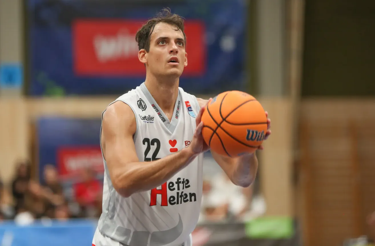 Ein Basketballspieler in einem weißen Trikot mit der Nummer 22 hält einen Ball. Er befindet sich auf einem Basketballplatz mit einer roten Wand im Hintergrund.
