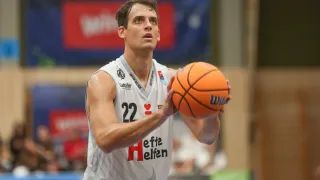 Ein Basketballspieler in einem weißen Trikot mit der Nummer 22 hält einen Ball. Er befindet sich auf einem Basketballplatz mit einer roten Wand im Hintergrund.
