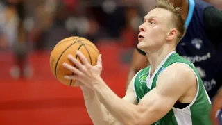 Ein Basketballspieler hält einen Basketball in der Hand, bereit zum Werfen, während ein anderer Spieler hinter ihm steht. Der Spielfeldboden ist rot.