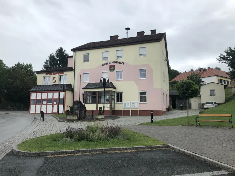 Ein Gebäude mit der Aufschrift 'Gemeindeamt' steht vor einer Feuerwache. Das Gebäude ist rosa mit einem braunen Dach und hat mehrere Fenster. Vor dem Gebäude stehen Pflanzen und ein Laternenpfahl.