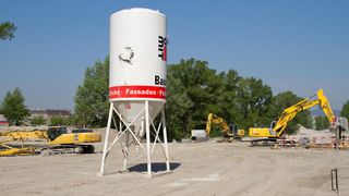 Ein großer weißer Silo steht auf einer Baustelle mit einigen gelben Baumaschinen in der Nähe. Auf dem Silo sind die Wörter 'Bau' und 'Fassaden' zu lesen. Bäume und ein Gebäude sind im Hintergrund sichtbar.