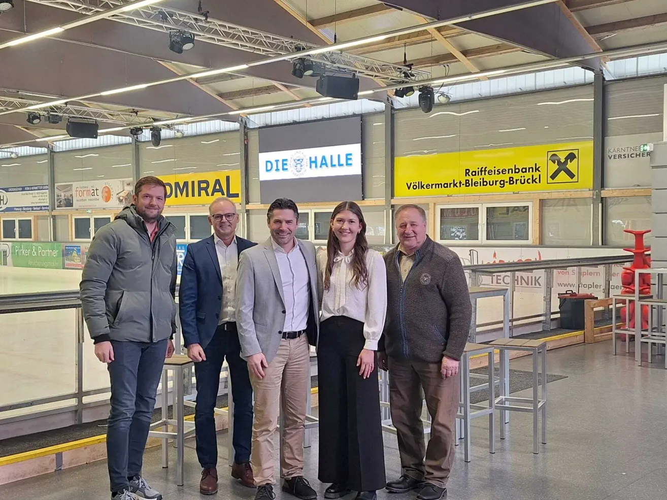 Fünf Personen stehen in einer Halle und lächeln für ein Foto. Hinter ihnen befindet sich ein gelbes Schild mit dem Wort Raiffeisenbank. Auf beiden Seiten der Halle befinden sich zwei Banner.