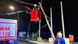 Feuerwehrleute arbeiten nachts an einem beschädigten Strommast, einer in einer Korbbühne und andere beobachten. Ein Feuerwehrwagen ist in der Nähe geparkt.
