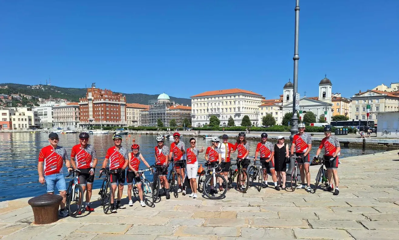Eine Gruppe von Radfahrern in passenden roten und weißen Uniformen steht am Wasser, mit Gebäuden und einem klaren Himmel im Hintergrund.