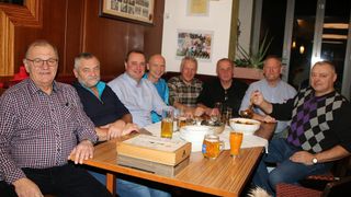 Sieben Männer sitzen lächelnd an einem Tisch in einem Restaurant. Der Tisch hat Biergläser, Teller und eine Schüssel. Die Männer tragen Freizeitkleidung. Die Wand hat Bilderrahmen und eine Topfpflanze.