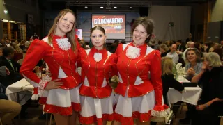 Drei Frauen in roten und weißen Uniformen stehen zusammen in einem Raum mit einer Menschenmenge im Hintergrund. Eine große Leinwand zeigt 'GILDENABEND'.