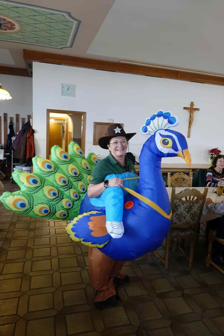 An older woman wearing a peacock costume sits on a chair in a dining area with a cross on the wall.