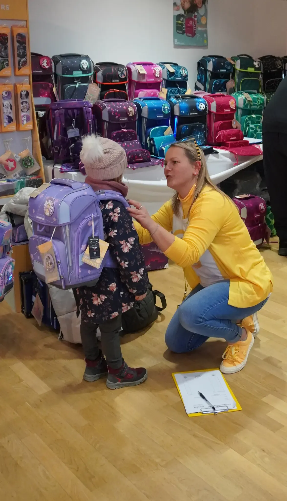 A woman is helping a child with a purple backpack in a store with colorful bags on shelves.