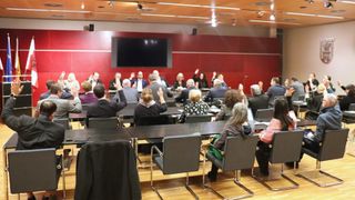 Eine Gruppe von Menschen sitzt in einem Raum auf Stühlen, wobei einige die Hände heben. Ein langer Tisch steht vor ihnen. Ein Monitor ist an der Wand hinter ihnen montiert. Einige Leute sitzen auf Stühlen auf der rechten Seite. Der Raum hat einen Holzboden und eine rote Wand.