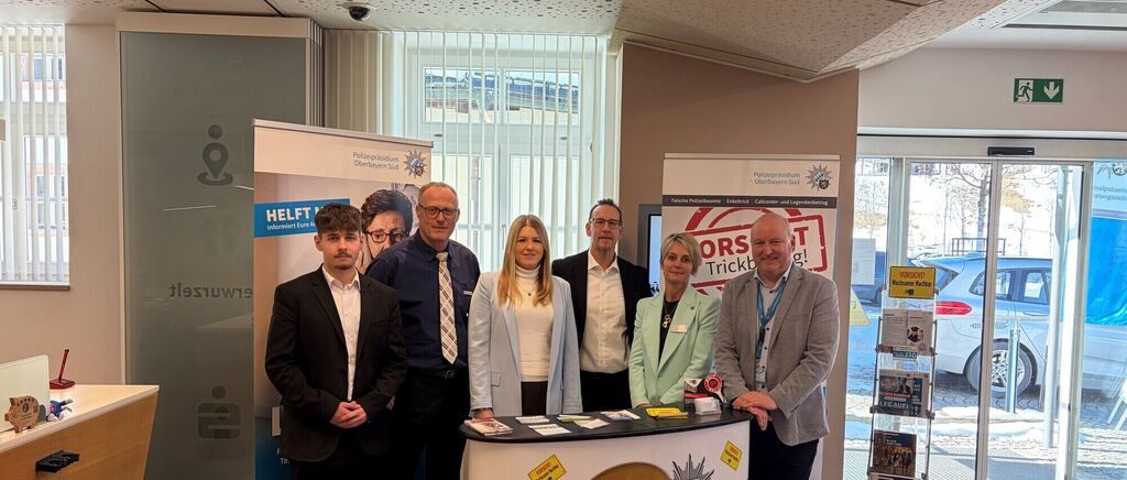 A group of people, including men and women, stand at a counter in a room with banners and books. One banner says 'HELFT' and another says 'ORST'. Outside the window, cars are parked.