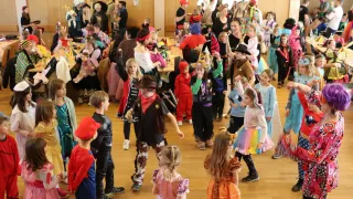 Kinder in verschiedenen Kostümen, darunter Piraten, Hexen und Tiere, tanzen in einem Raum mit Holzboden. Einige von ihnen halten Puppen und Spielzeug. Im Hintergrund sitzen Menschen an einem Tisch mit Essen und Getränken.