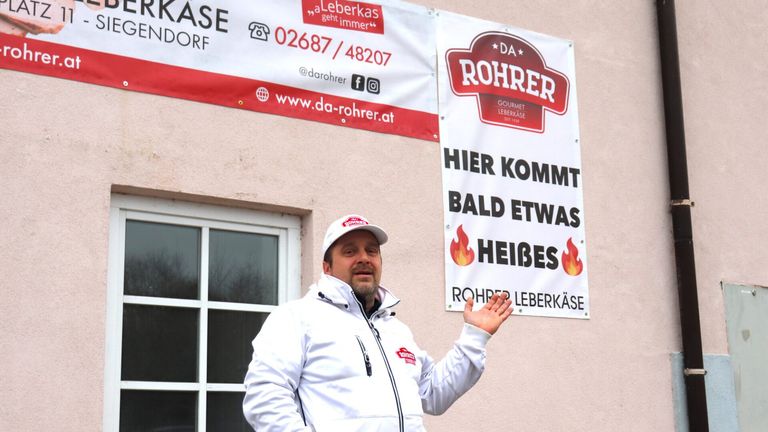 A man in a white jacket and cap stands in front of a building with a banner that says Rohrer and Hier Kommt Bald Etwas Heibes.