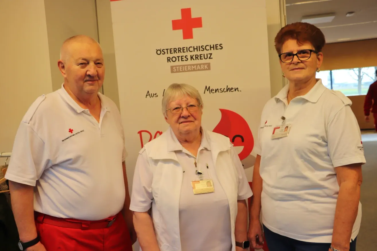 Drei Personen stehen vor einem Banner mit einem roten Kreuz und den Worten 'Aus I Menschen'. Die beiden Frauen tragen Brillen und Ausweise. Der Mann trägt einen roten Gürtel.