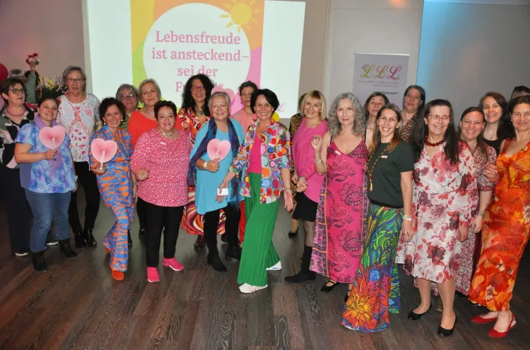 A group of women in vibrant dresses, smiling and posing for a photo. They hold pink fans and stand in front of a projector screen with the text 'Lebensfreude ist ansteckend-sei der F.' Behind them, there is a banner with the text 'LLL'.