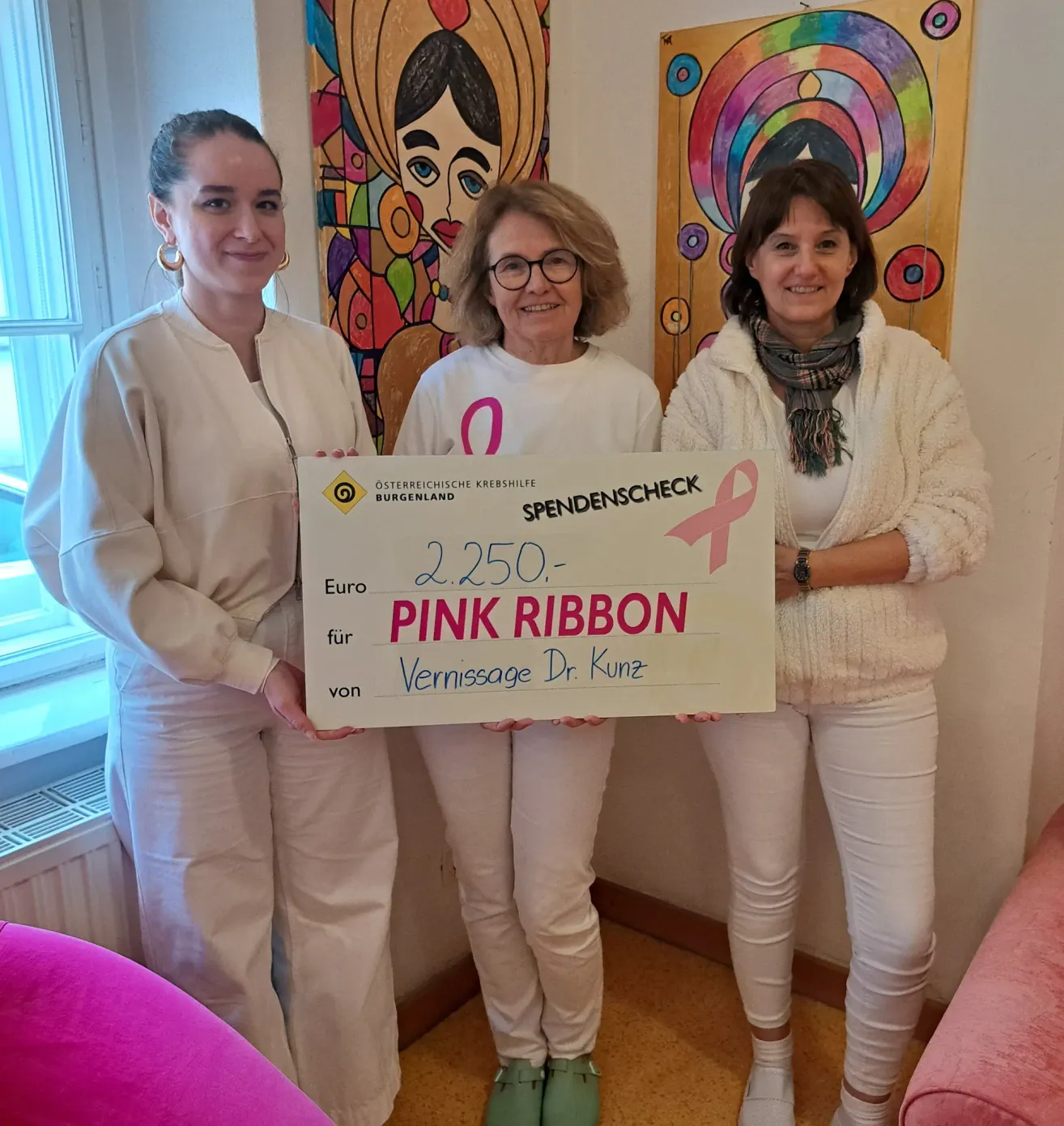 Three women hold a donation check for Pink Ribbon. The middle woman wears glasses. They stand in front of two large paintings. The left painting depicts a woman, and the right one has a colorful design.