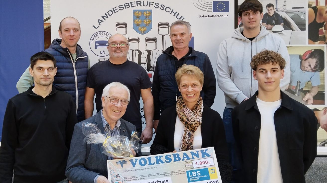 Several individuals stand together, smiling, holding a check from Volksbank for 1,800 euros. They are in front of a wall with a logo and text that reads Landesberufsschule.