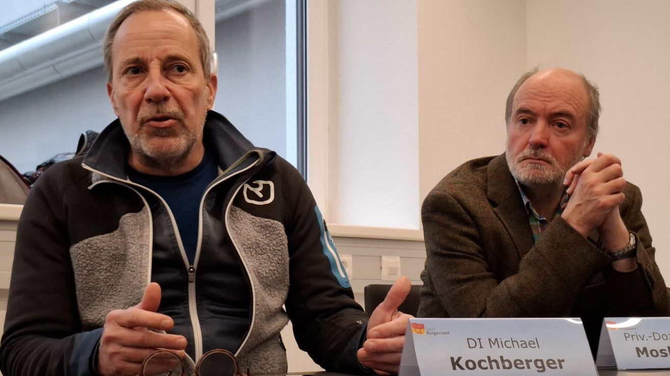 Two men sit facing each other in a room. The man on the left wears a jacket with a logo and speaks, gesturing. The man on the right, with a beard, listens attentively. A nameplate with 'DI Michael Kochbe' is visible.