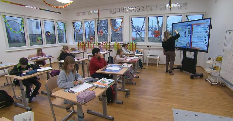 A classroom with several students sitting at desks. A teacher stands before a large screen, explaining something. There are large windows and decorations on the walls.