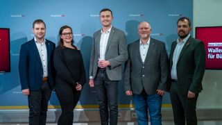 Vier Personen stehen vor einer blauen Wand mit dem Text Burgenlandpur. Die Frau auf der linken und der Mann auf der rechten Seite lächeln. Der Mann in der Mitte hält ein Glas.